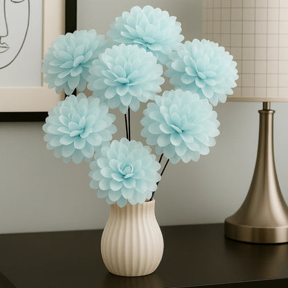 Light blue flowers in a white vase on a dark surface with a lamp and framed picture in the background.