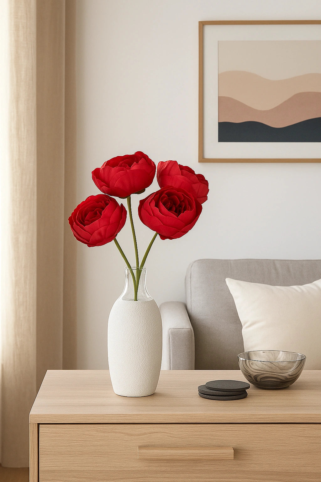 Red flowers in a white vase on a wooden side table with a sofa and wall art in the background.