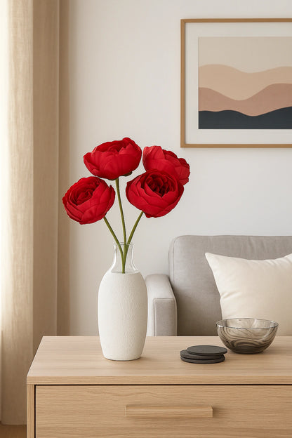 Red flowers in a white vase on a wooden side table with a sofa and wall art in the background.